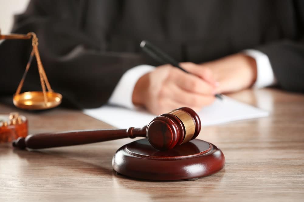 Image is of a judge writing legal documents with a gavel and scales of justice in the foreground, concept of an alimony attorney representing clients during court proceedings involving spousal support.