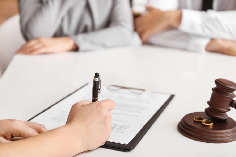 Image is of a person signing legal documents while a couple sits nearby, concept of an annulment attorney preparing and reviewing annulment paperwork.