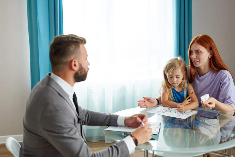 Image is of a parent meeting with a lawyer while a child colors at the table, concept of a child custody attorney discussing parenting arrangements.