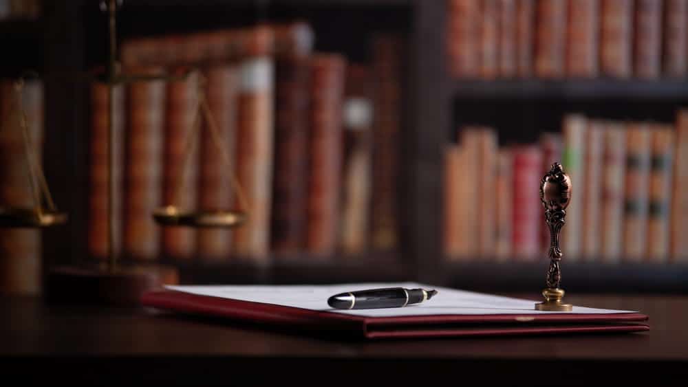 Image is of legal documents and a pen on a desk with law books in the background, concept of child support attorney.