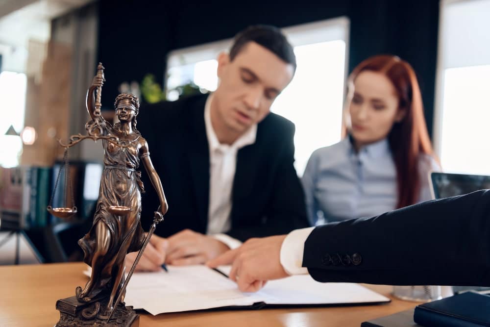 Image is of an attorney meeting with a couple to review legal documents beside a Lady Justice statue, concept of a property division attorney helping resolve marital asset disputes.
