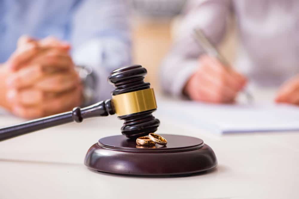 Image is of a judge's gavel with wedding rings while a couple signs legal documents in the background, concept of a prenuptial agreement attorney assisting with premarital contract preparation.