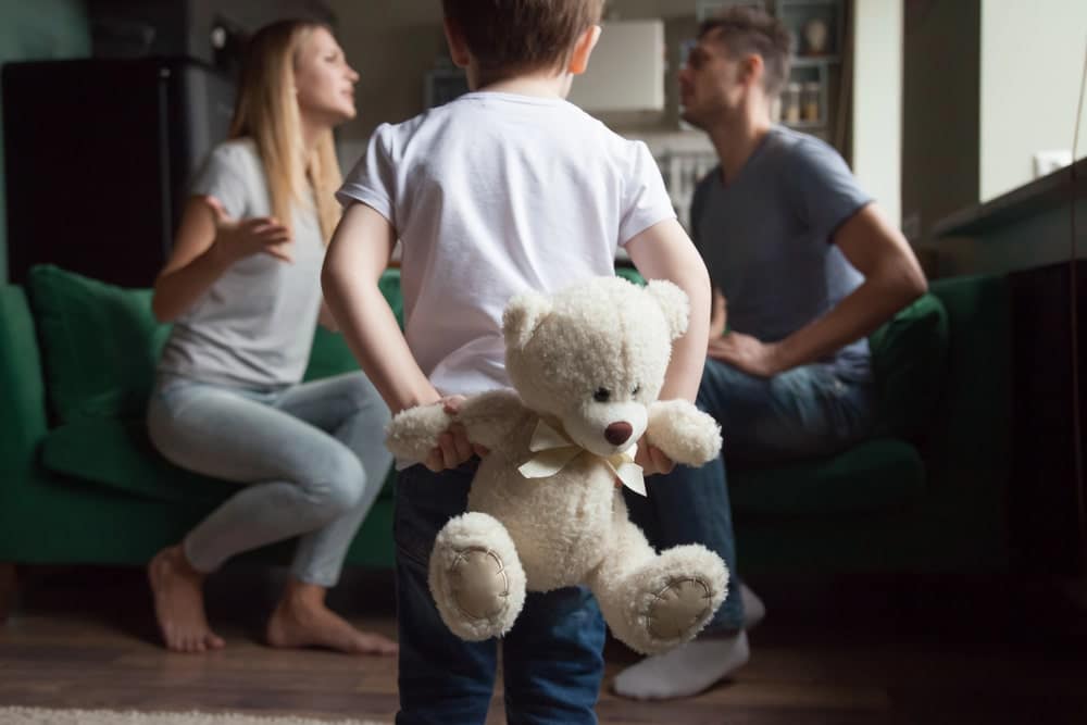Image is of a child holding a teddy bear while parents argue nearby, concept of divorce with children affecting a child's emotional well-being.
