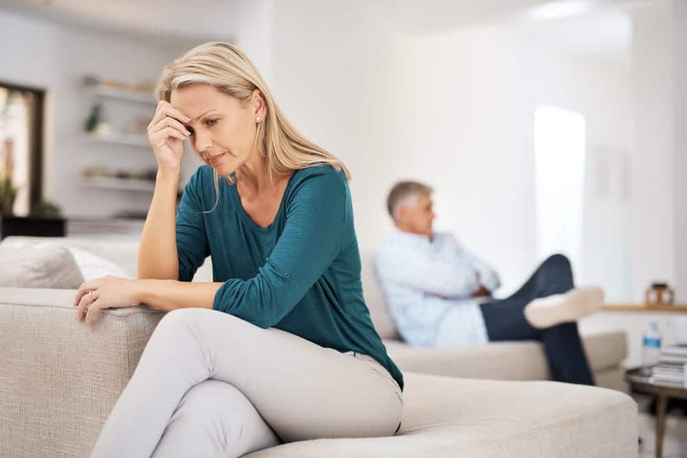 Image is of a distressed spouse sitting apart from a partner at home, concept of an alienation of affection attorney helping with marital interference claims.