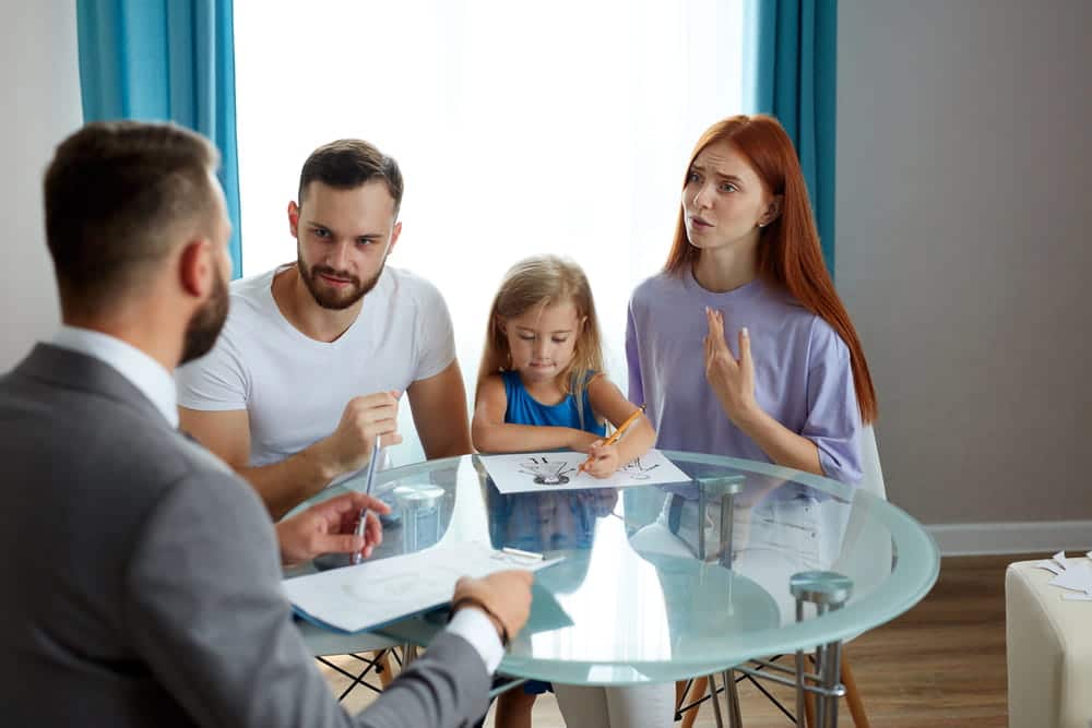 Image is of parents discussing family arrangements with a professional while their child sits nearby, representing joint custody decisions.